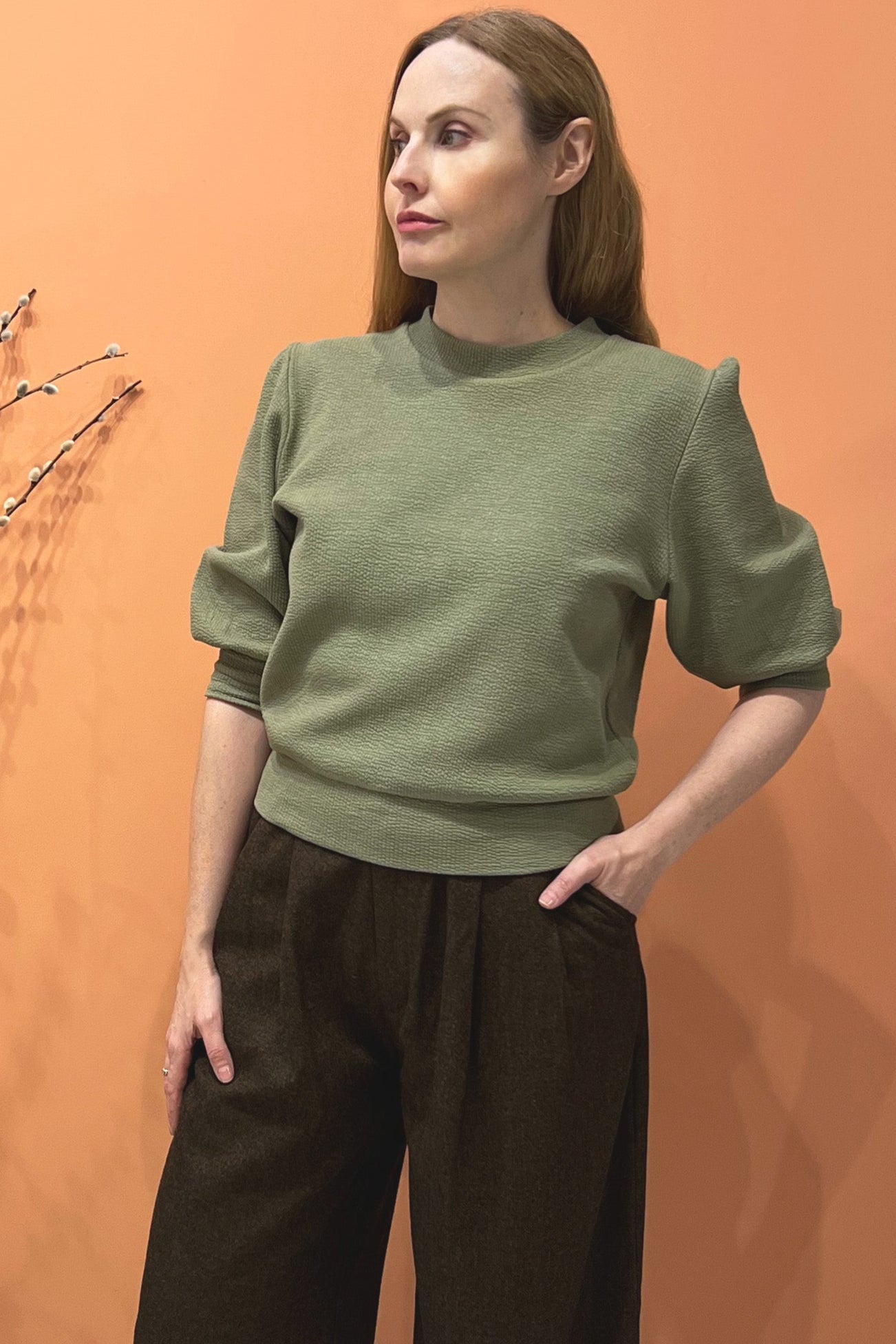 A woman wearing the Bridge Sweater by Allison Wonderland in Matcha, featuring elbow length sleeves and a ribbed neckline, cuff, and hem. She's wearing it with the Memory Pants in Herringbone and standing in front of an orange wall. 