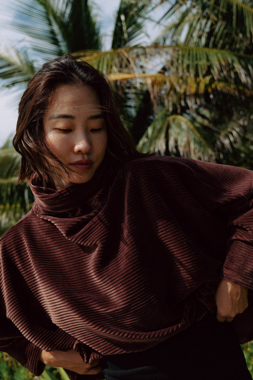 A woman wearing the Amsterdam Sweater by MAS in Cacao, an oversized ribbed velvet sweater with a chimney neckline and long batwing sleeves. 