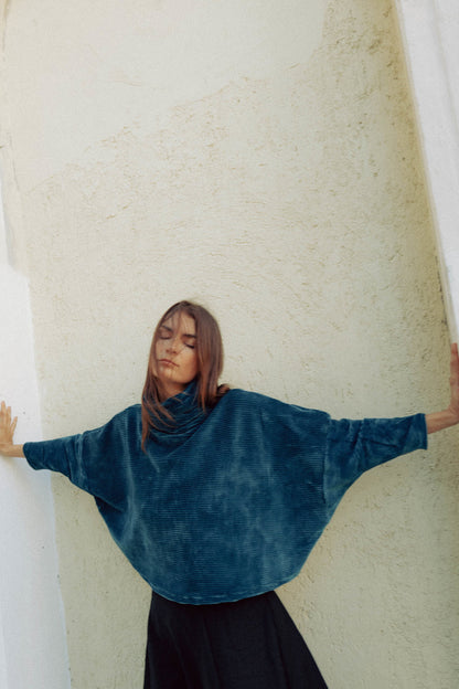 A woman wearing the Amsterdam Sweater by MAS in Juniper, an oversized ribbed velvet sweater with a chimney neckline and long batwing sleeves. She is wearing it with a black skirt pant and standing against a white wall. 