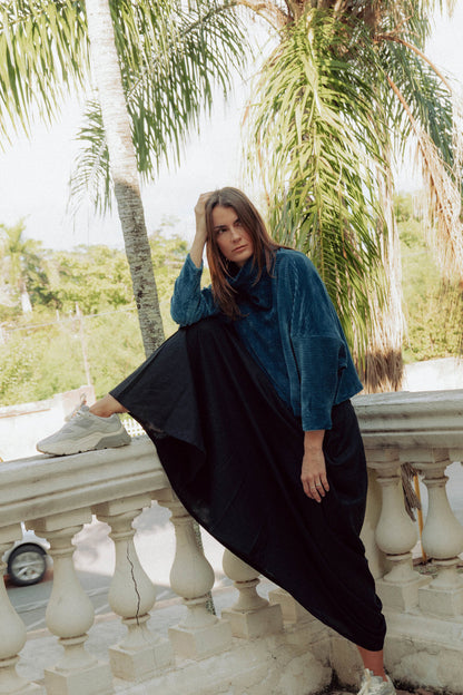 A woman wearing the Amsterdam Sweater by MAS in Juniper, an oversized ribbed velvet sweater with a chimney neckline and long batwing sleeves. She is wearing it with a black skirt and sitting on a balcony.