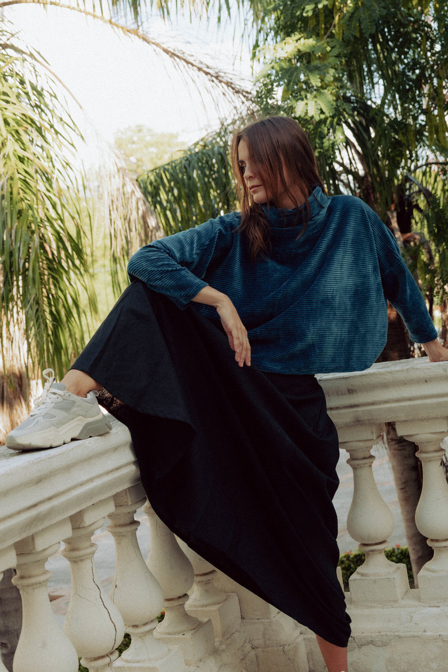A woman wearing the Amsterdam Sweater by MAS in Juniper, an oversized ribbed velvet sweater with a chimney neckline and long batwing sleeves. She is wearing it with a black skirt and sitting on a balcony.