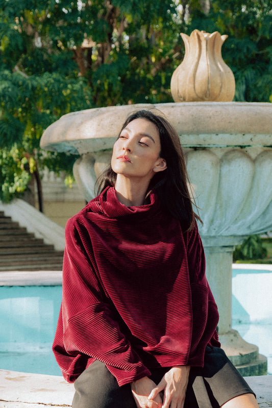 A woman wearing the Amsterdam Sweater by MAS in Rosewood, a an oversized ribbed velvet sweater with a chimney neckline and long batwing sleeves. She is sitting by a fountain.