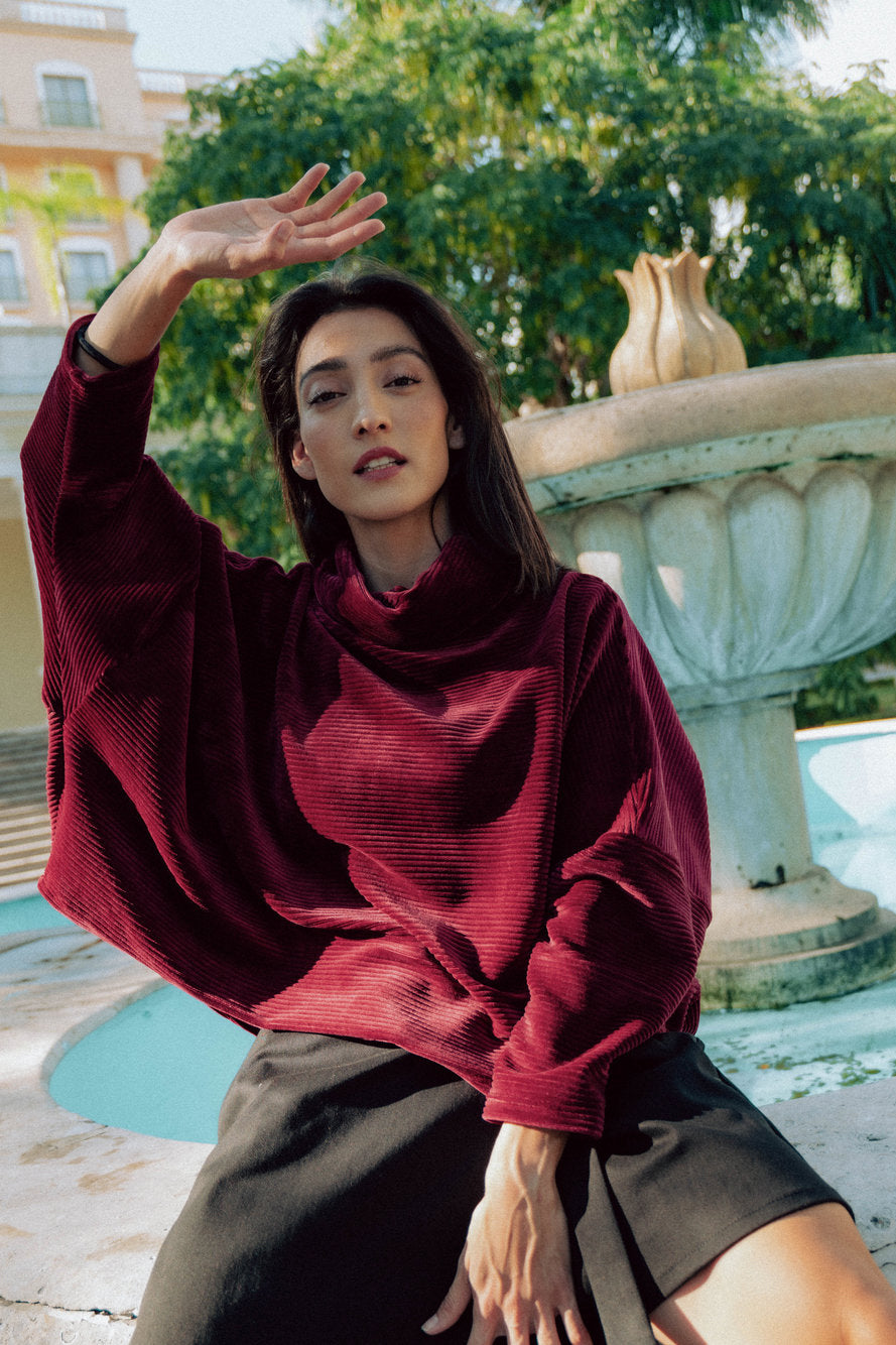 A woman wearing the Amsterdam Sweater by MAS in Rosewood, a an oversized ribbed velvet sweater with a chimney neckline and long batwing sleeves. She is wearing it with a black skirt and sitting by a fountain.