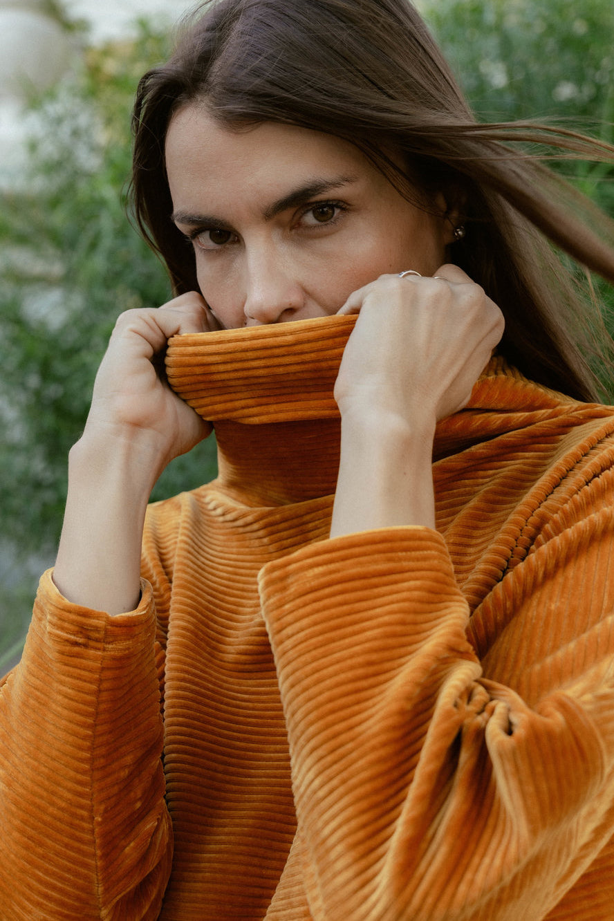 A woman wearing the Amsterdam Sweater by MAS in Tumeric, an oversized ribbed velvet sweater with a chimney neckline and long batwing sleeves.