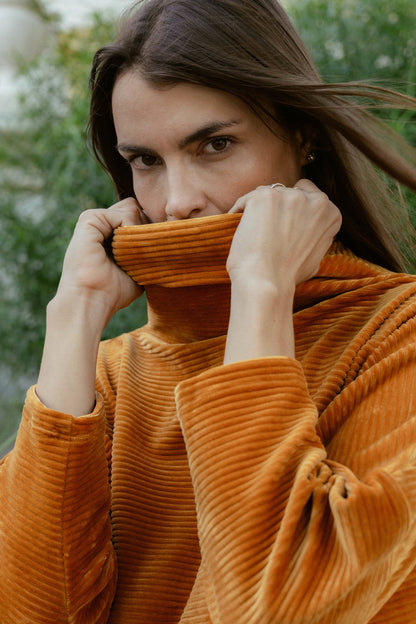 A woman wearing the Amsterdam Sweater by MAS in Tumeric, an oversized ribbed velvet sweater with a chimney neckline and long batwing sleeves.
