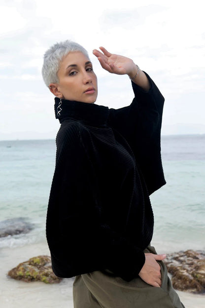 A woman wearing the Amsterdam Sweater by MAS in Pepper, an oversized ribbed velvet sweater with a chimney neckline and long batwing sleeves. She is wearing it with green pants and standing on a beach. 