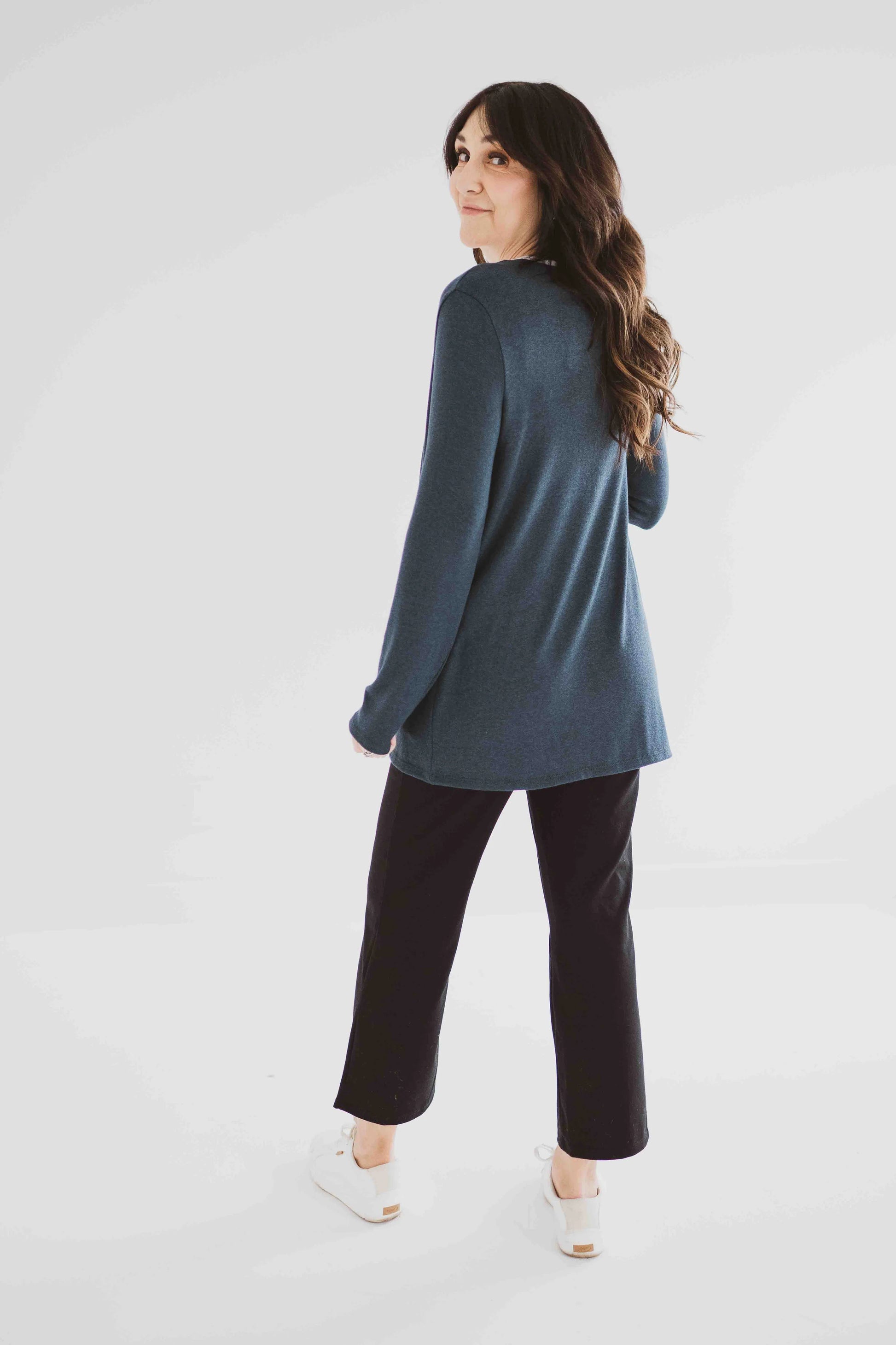 Back view of a woman wearing the Carolina Cardigan by Ang Hill in Juniper, an open cardigan with long sleeves. She is wearing it with black pants and a white top and is standing in front of a white background. 