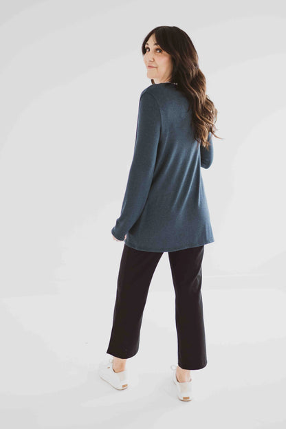 Back view of a woman wearing the Carolina Cardigan by Ang Hill in Juniper, an open cardigan with long sleeves. She is wearing it with black pants and a white top and is standing in front of a white background. 