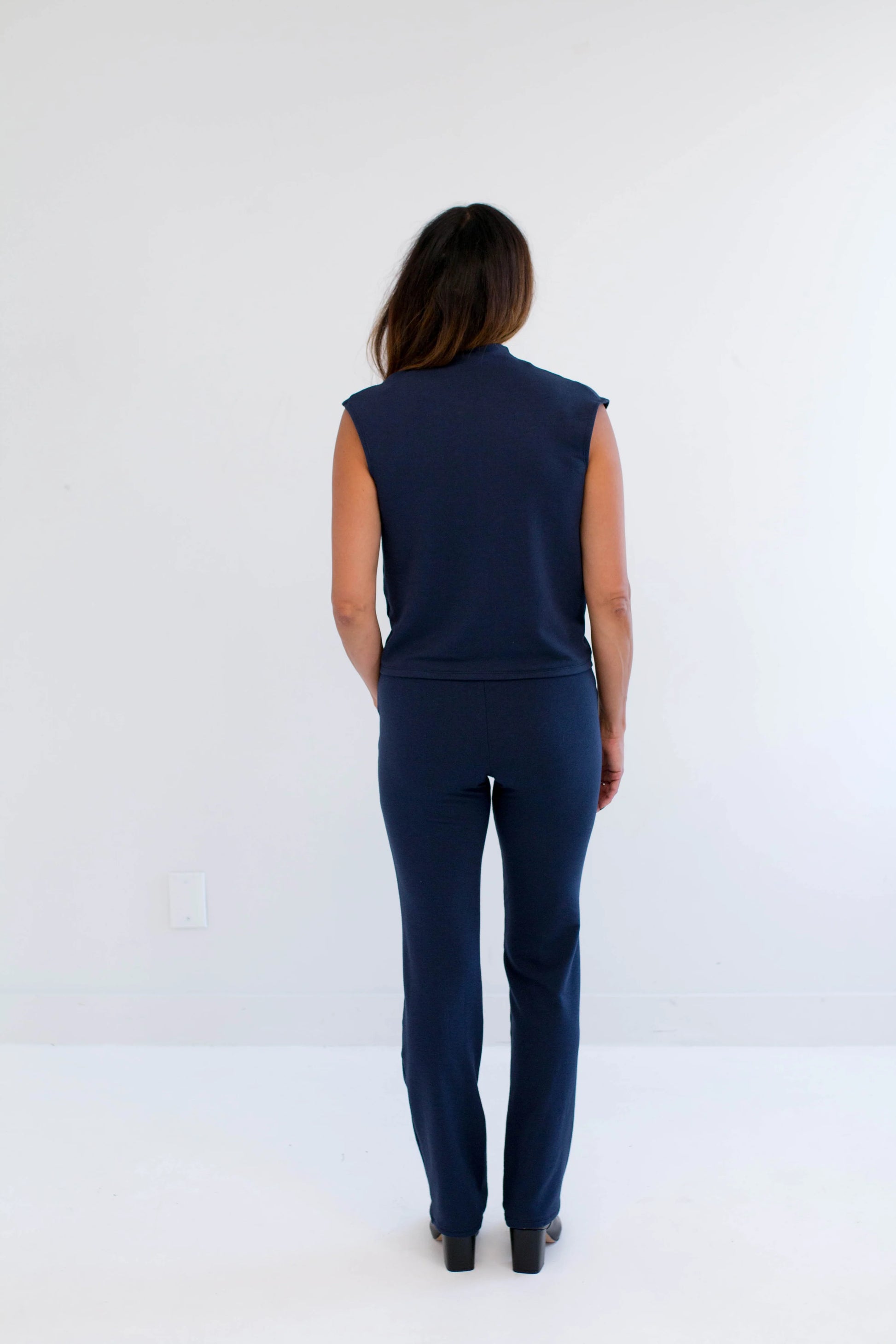 Back view of a woman wearing the Dori Mockneck navy sleeveless top from Ang Hill, standing against a white background