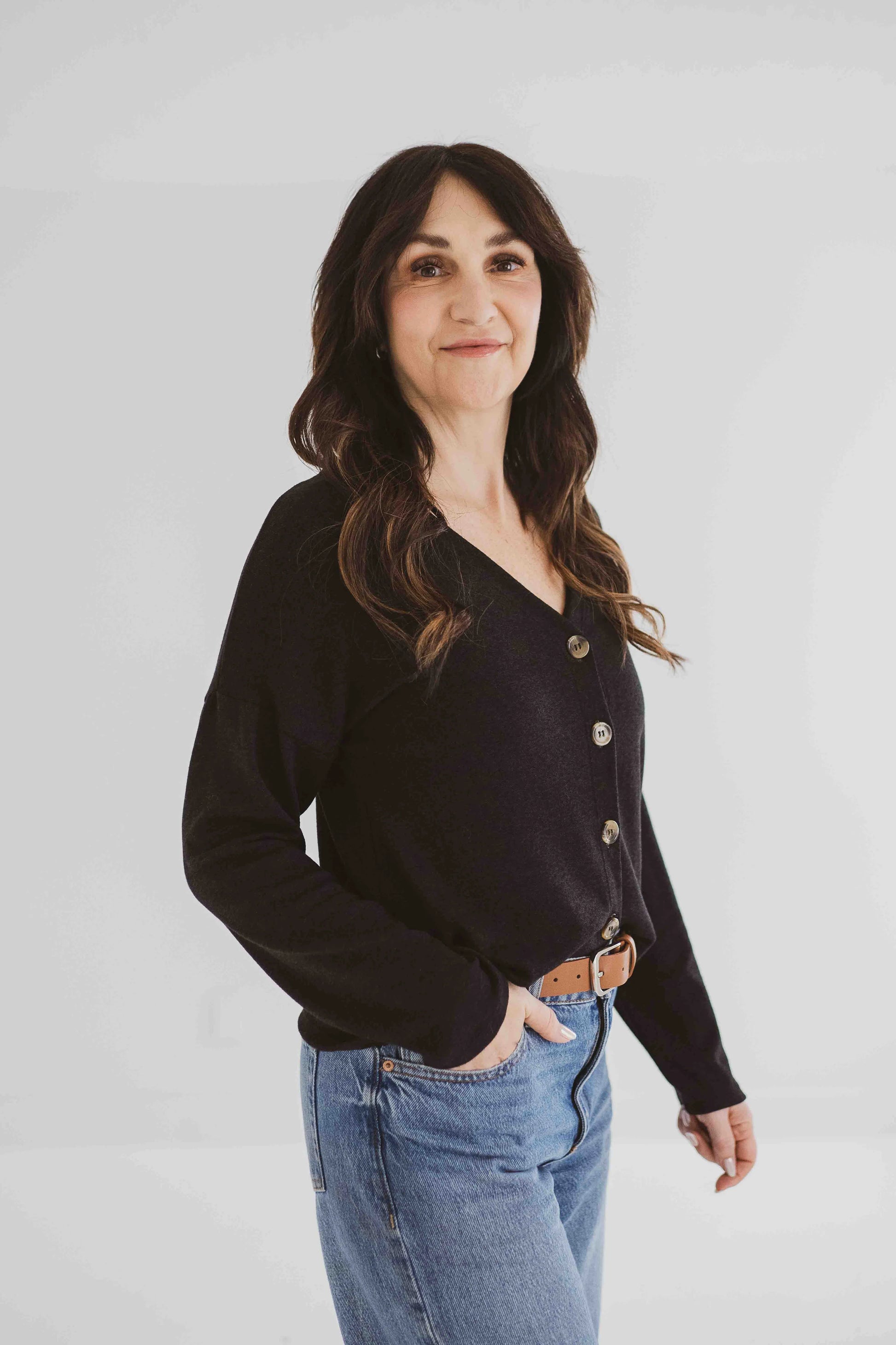 A woman wearing the Holly Cropped Cardigan by Ang Hill in Black, a v-neck cardigan with drop sleeves and tortoiseshell buttons. She is wearing it with jeans and is standing in front of a white background. 