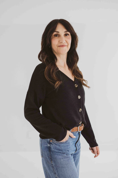 A woman wearing the Holly Cropped Cardigan by Ang Hill in Black, a v-neck cardigan with drop sleeves and tortoiseshell buttons. She is wearing it with jeans and is standing in front of a white background. 