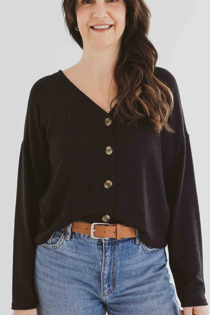 A woman wearing the Holly Cropped Cardigan by Ang Hill in Black, a v-neck cardigan with drop sleeves and tortoiseshell buttons. She is wearing it with jeans and is standing in front of a white background. 