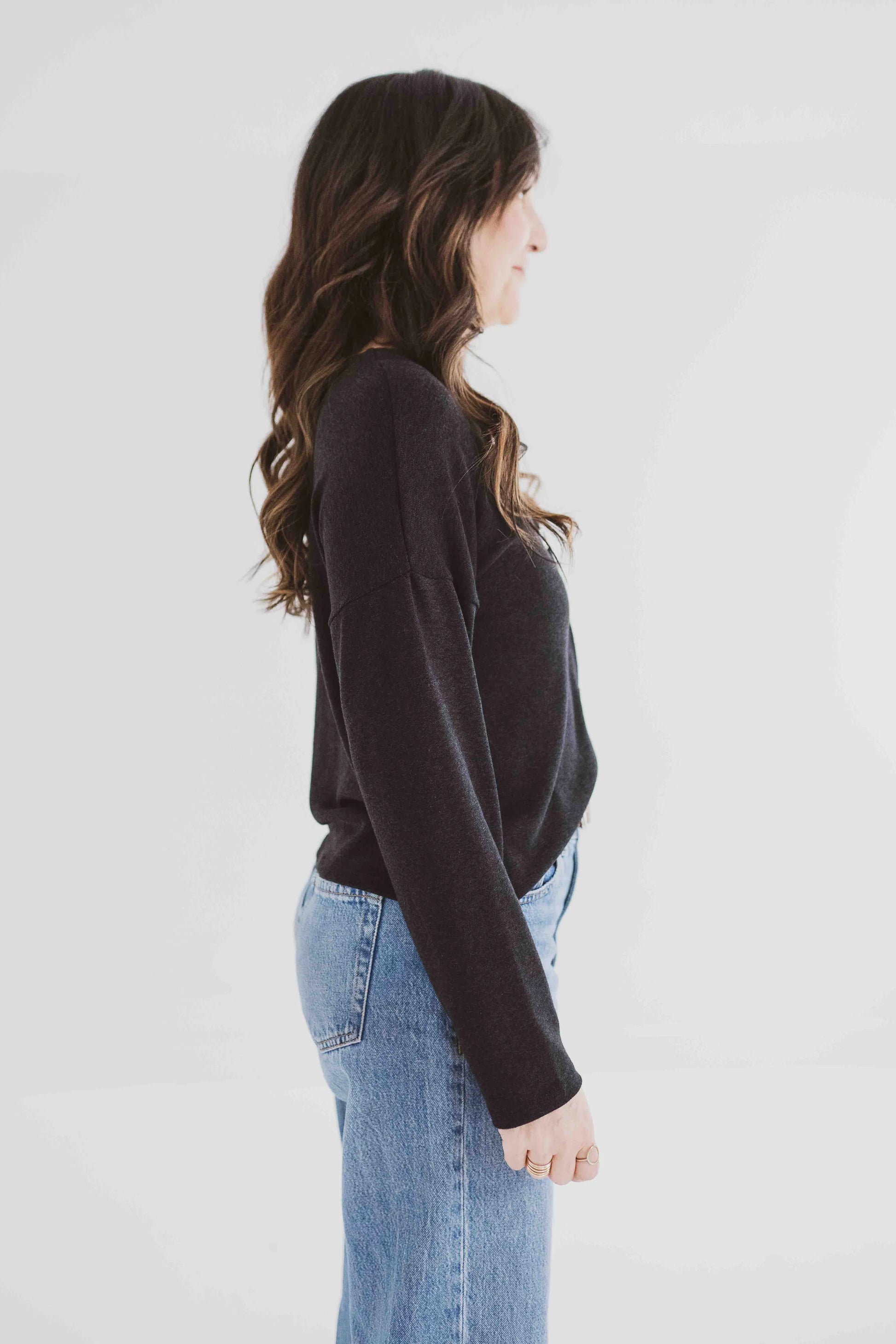 Side view of a woman wearing the Holly Cropped Cardigan by Ang Hill in Black, a v-neck cardigan with drop sleeves and tortoiseshell buttons. She is wearing it with jeans and is standing in front of a white background. 