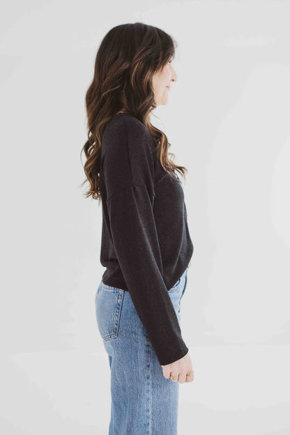 Side view of a woman wearing the Holly Cropped Cardigan by Ang Hill in Black, a v-neck cardigan with drop sleeves and tortoiseshell buttons. She is wearing it with jeans and is standing in front of a white background. 