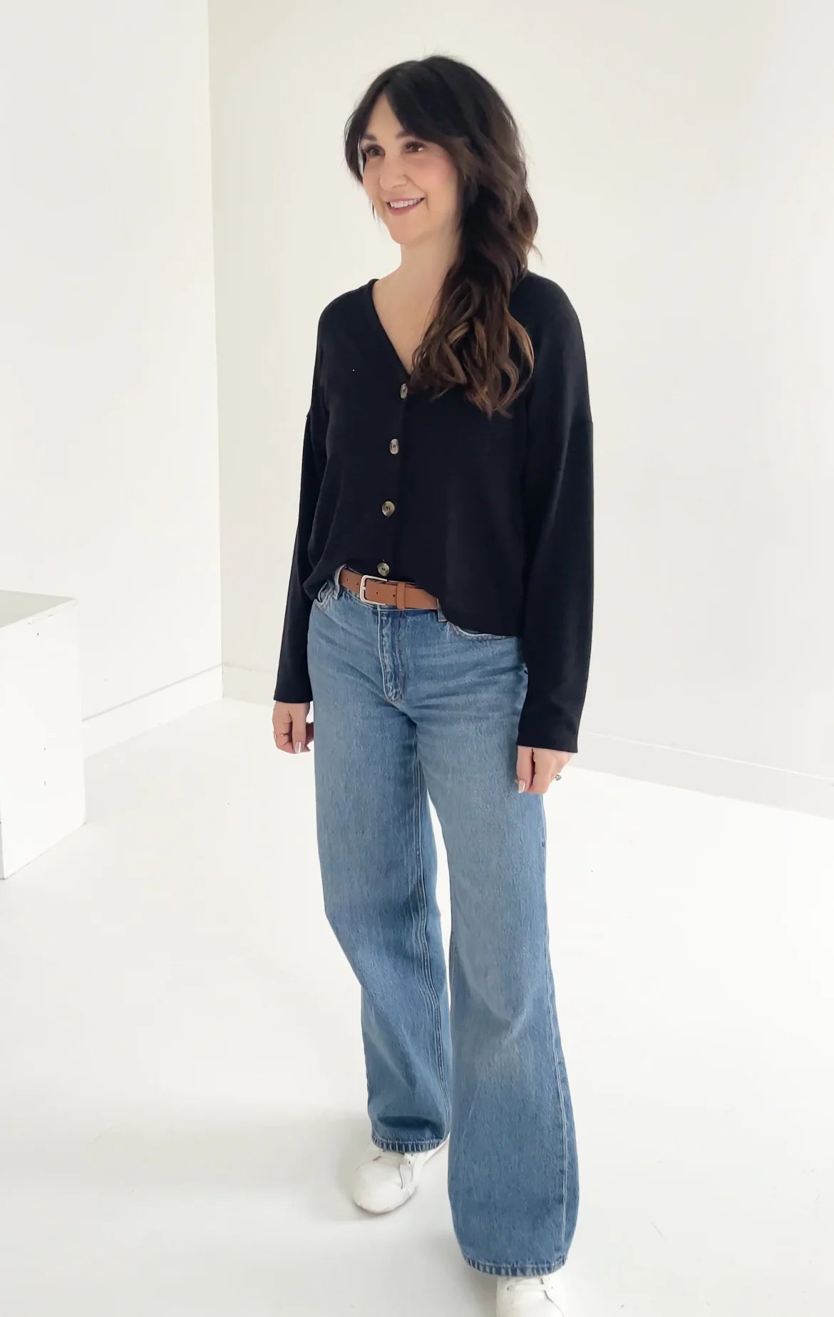 A woman wearing the Holly Cropped Cardigan by Ang Hill in Black, a v-neck cardigan with drop sleeves and tortoiseshell buttons. She is wearing it with jeans and is standing in front of a white background. 