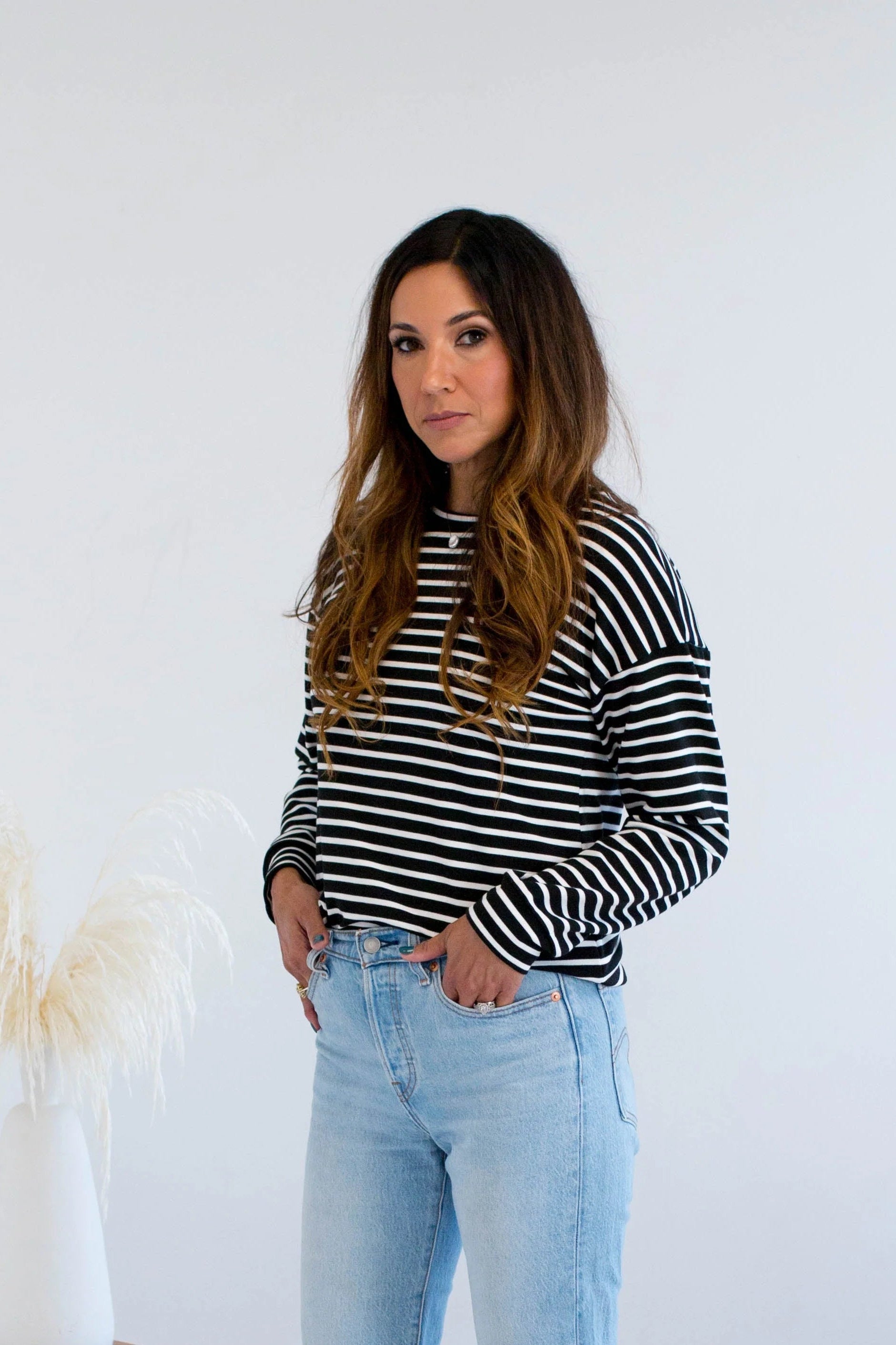 A woman wearing the Munro Bamboo Crewneck sweater by Ang Hill in black and white stripes. She is wearing it with light blue jeans against a white background.