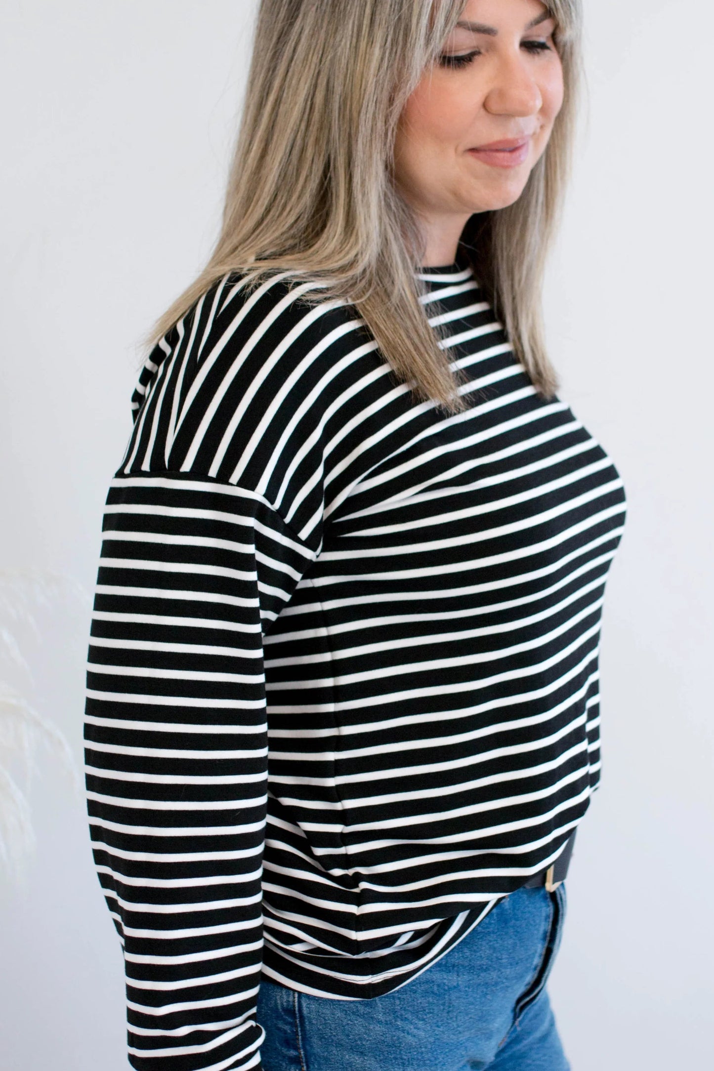 Side view of a woman wearing the Munro Bamboo Crewneck sweater by Ang Hill in black and white stripes. She is wearing it with light blue jeans against a white background.