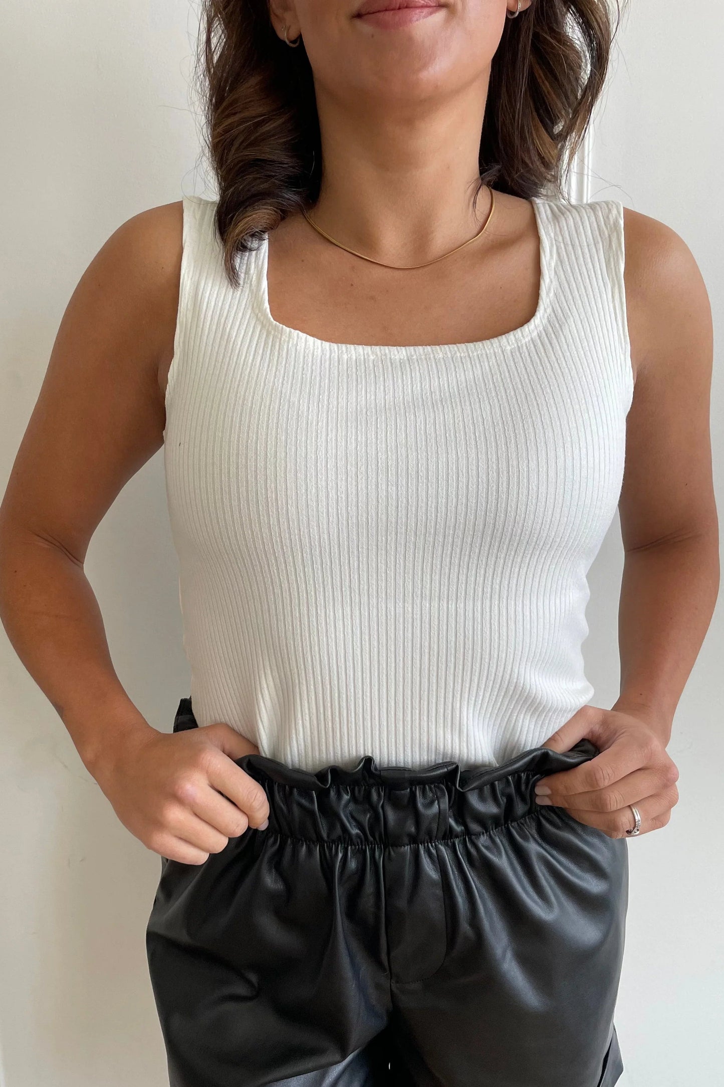 Close up of a woman wearing the Ang Hill Tracey Tank in Vanilla, with wide straps, ribbed fabric, and a square neckline. She is wearing it with black shorts and standing against a white paneled wall.