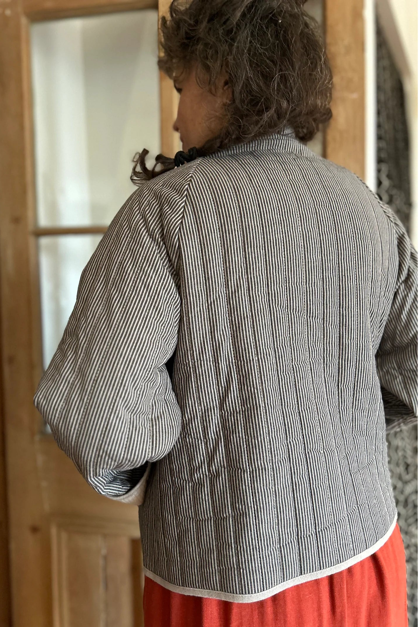 Back view of a woman wearing the Artemesia Jacket by Kazak in Taupe Stripe, a quilted jacket with snaps up the front and large patch pockets. She is wearing it over  the Lavendula Dress Paprika and standing in front of a wood and glass door. 