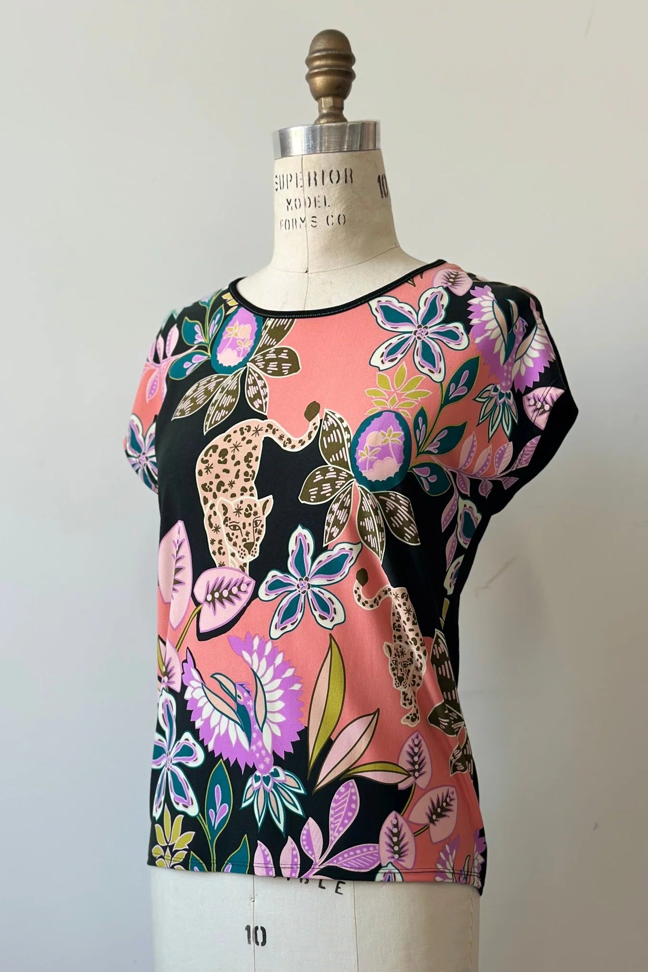 The Badia T-shirt by Kollontai in Bubblegum, featuring a patterned front with leopards, flowers and birds and a solid black back with cap sleeves and a hi-low hem, is shown on a mannequin in front of a white background.