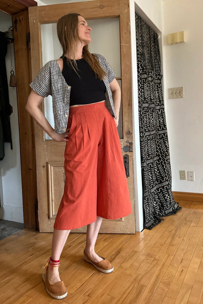 A woman wearing the Petunia Pants by Kazak in Cayenne, wide legged cropped pants with a waistband that's smooth in front but elastic at the back, front pleats, and pockets. She is wearing them with a black tank and an open checkered shirt and standing in front of a wooden door. 