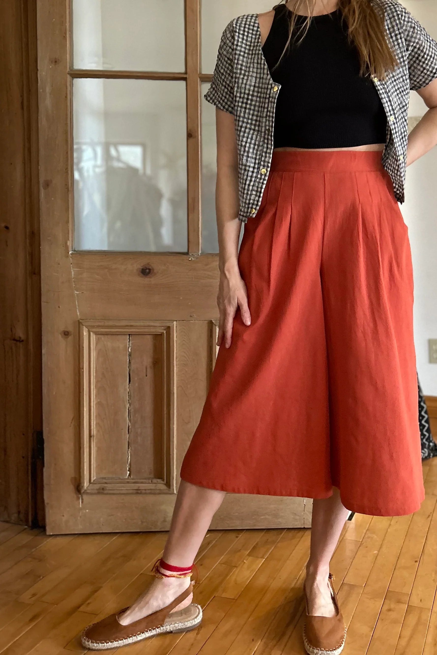 Neck down view of a woman wearing the Petunia Pants by Kazak in Cayenne, wide legged cropped pants with a waistband that's smooth in front but elastic at the back, front pleats, and pockets. She is wearing them with a black tank and an open checkered shirt and standing in front of a wooden door. 