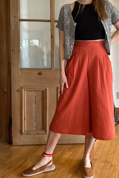 Neck down view of a woman wearing the Petunia Pants by Kazak in Cayenne, wide legged cropped pants with a waistband that's smooth in front but elastic at the back, front pleats, and pockets. She is wearing them with a black tank and an open checkered shirt and standing in front of a wooden door. 