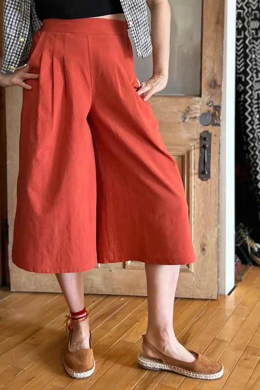Neck down view of a woman wearing the Petunia Pants by Kazak in Cayenne, wide legged cropped pants with a waistband that's smooth in front but elastic at the back, front pleats, and pockets. She is wearing them with a black tank and an open checkered shirt and standing in front of a wooden door. 