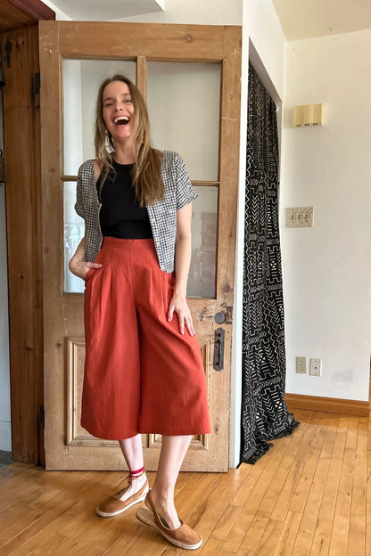 A woman wearing the Petunia Pants by Kazak in Cayenne, wide legged cropped pants with a waistband that's smooth in front but elastic at the back, front pleats, and pockets. She is wearing them with a black tank and an open checkered shirt and standing in front of a wooden door. 