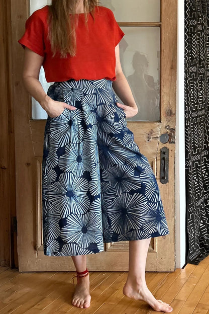 A woman wearing the Petunia Pants by Kazak in Indigo Floral,  wide legged cropped pants with a waistband that's smooth in front but elastic at the back, front pleats, and pockets. She is wearing them with an orange top and standing in front of a wooden door. 