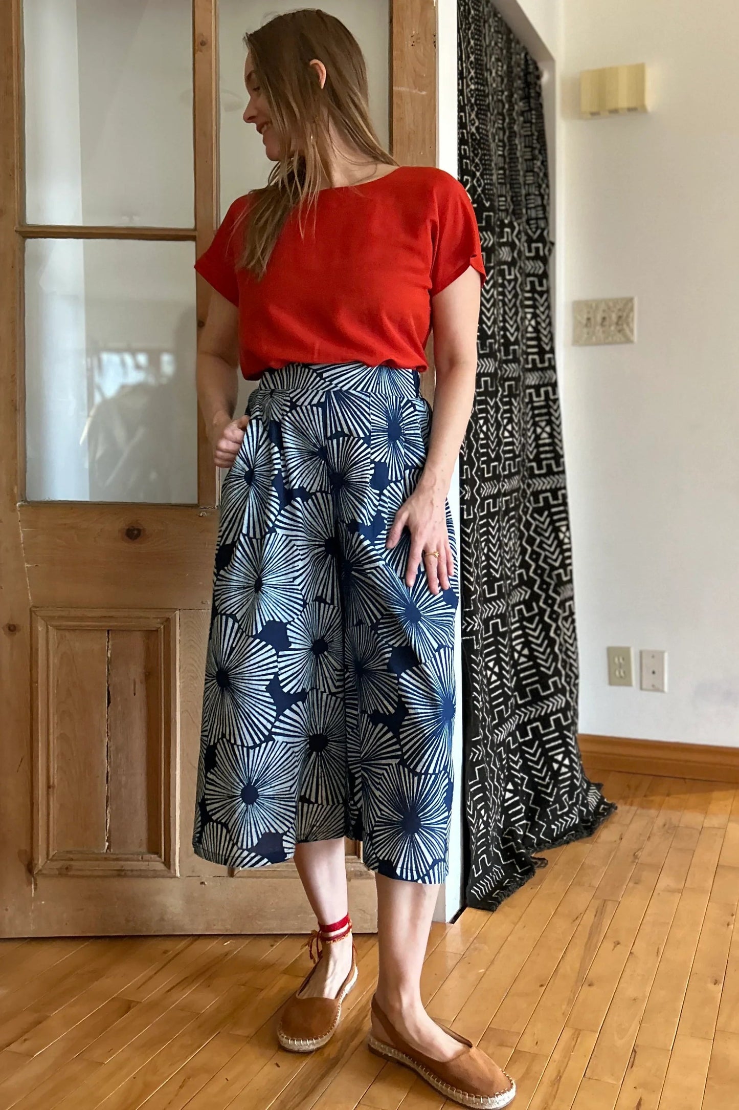 A woman wearing the Petunia Pants by Kazak in Indigo Floral,  wide legged cropped pants with a waistband that's smooth in front but elastic at the back, front pleats, and pockets. She is wearing them with an orange top and standing in front of a wooden door. 