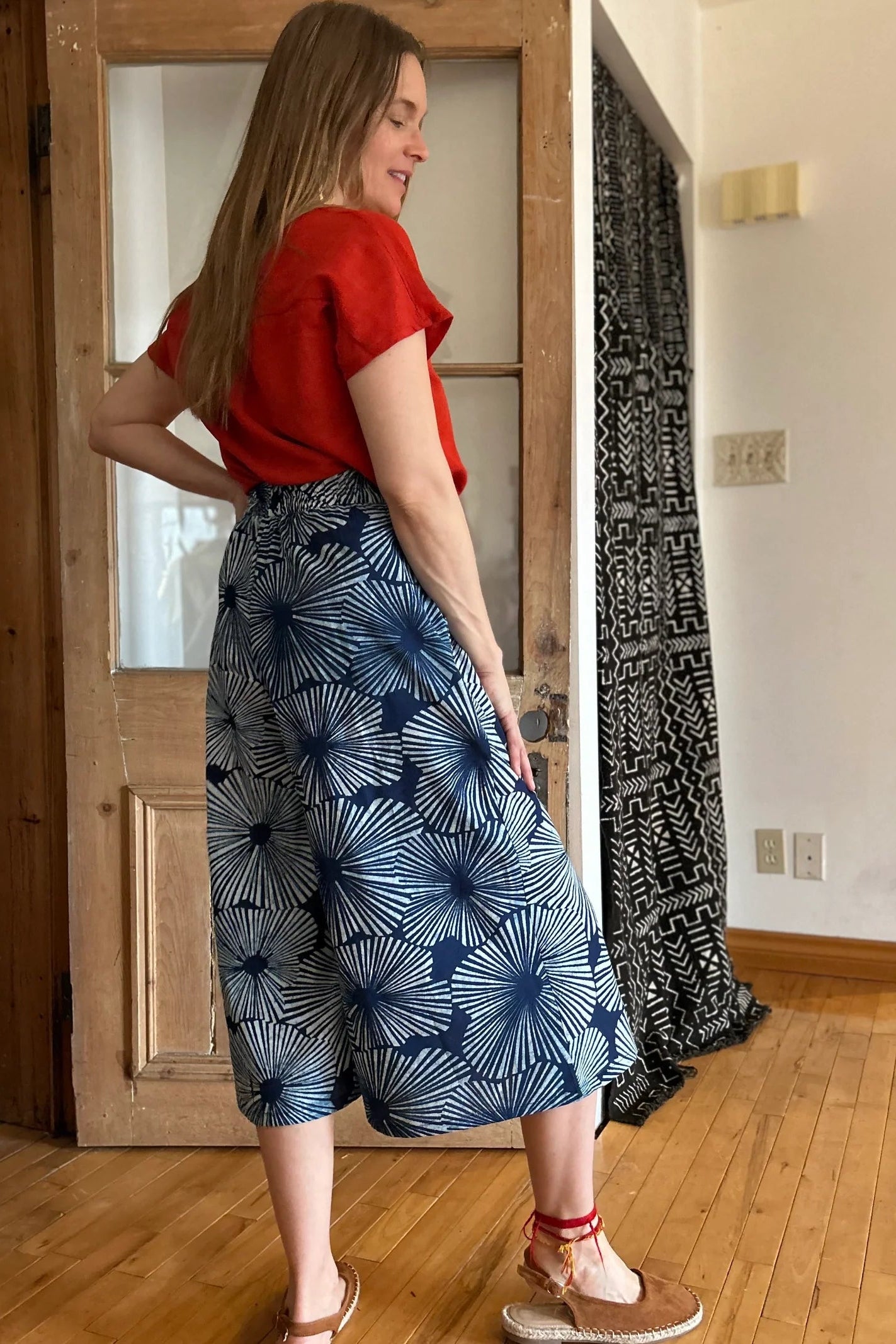 Side view of a woman wearing the Petunia Pants by Kazak in Indigo Floral,  wide legged cropped pants with a waistband that's smooth in front but elastic at the back, front pleats, and pockets. She is wearing them with an orange top and standing in front of a wooden door. 