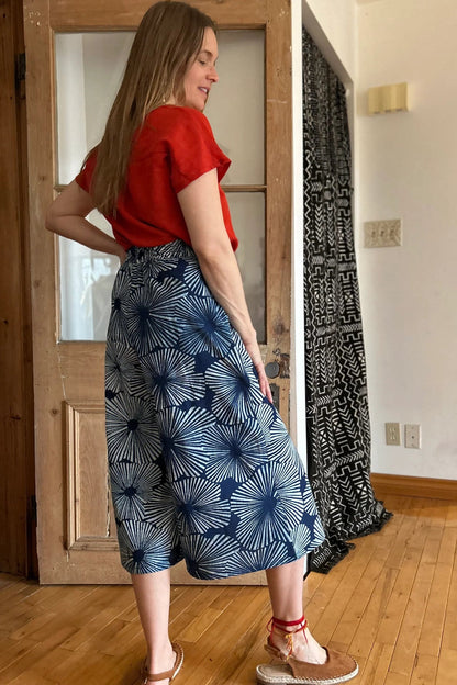 Side view of a woman wearing the Petunia Pants by Kazak in Indigo Floral,  wide legged cropped pants with a waistband that's smooth in front but elastic at the back, front pleats, and pockets. She is wearing them with an orange top and standing in front of a wooden door. 