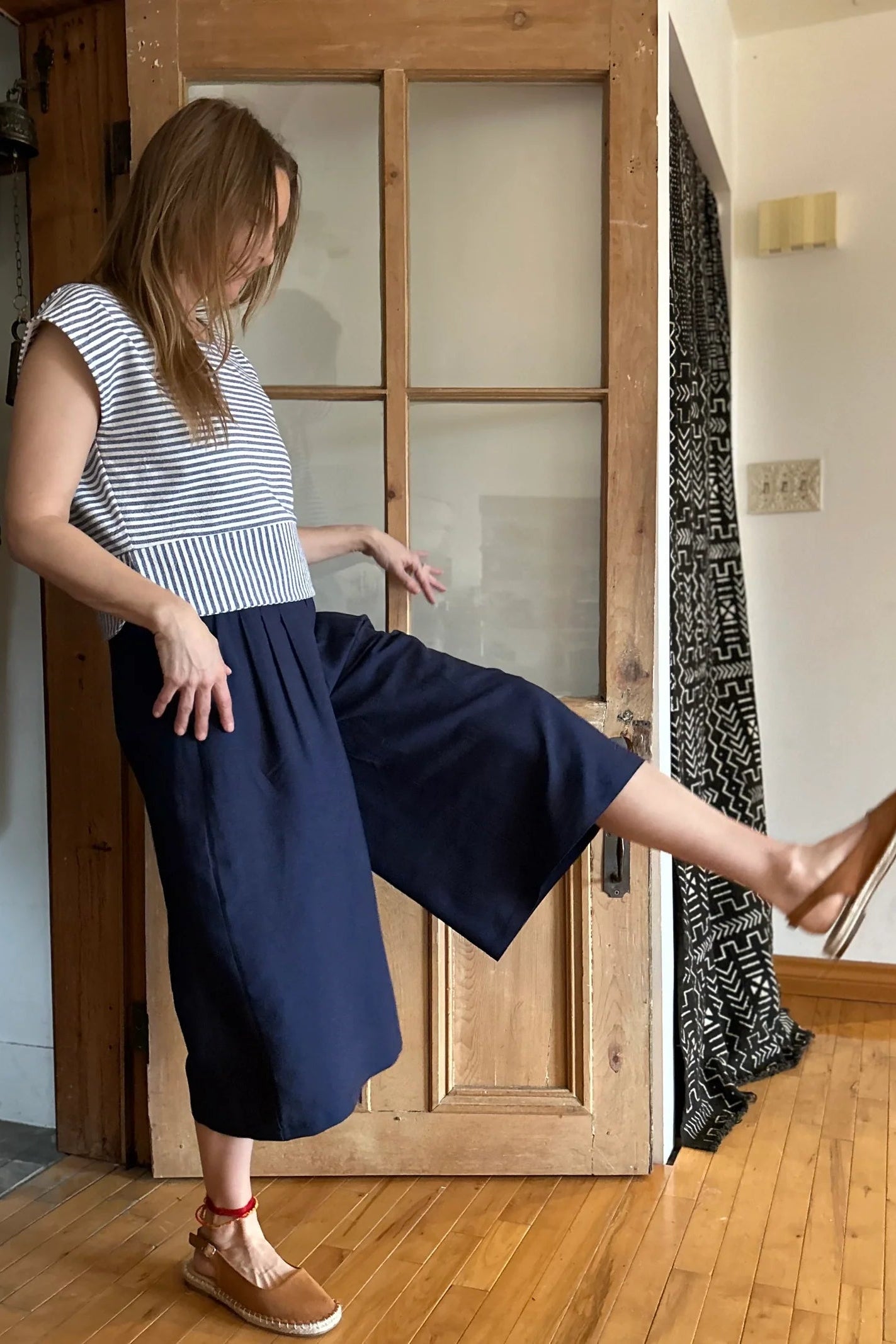 Side view of a woman wearing the Petunia Pants by Kazak in Indigo, wide legged cropped pants with a waistband that's smooth in front but elastic at the back, front pleats, and pockets. She is wearing them with the Tulipe top and standing in front of a wooden door. 