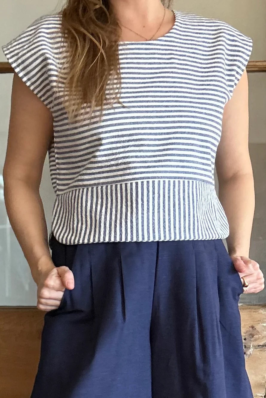 Torso view of a woman wearing the Petunia Pants by Kazak in Indigo, wide legged cropped pants with a waistband that's smooth in front but elastic at the back, front pleats, and pockets. She is wearing them with the Tulipe top and standing in front of a wooden door. 