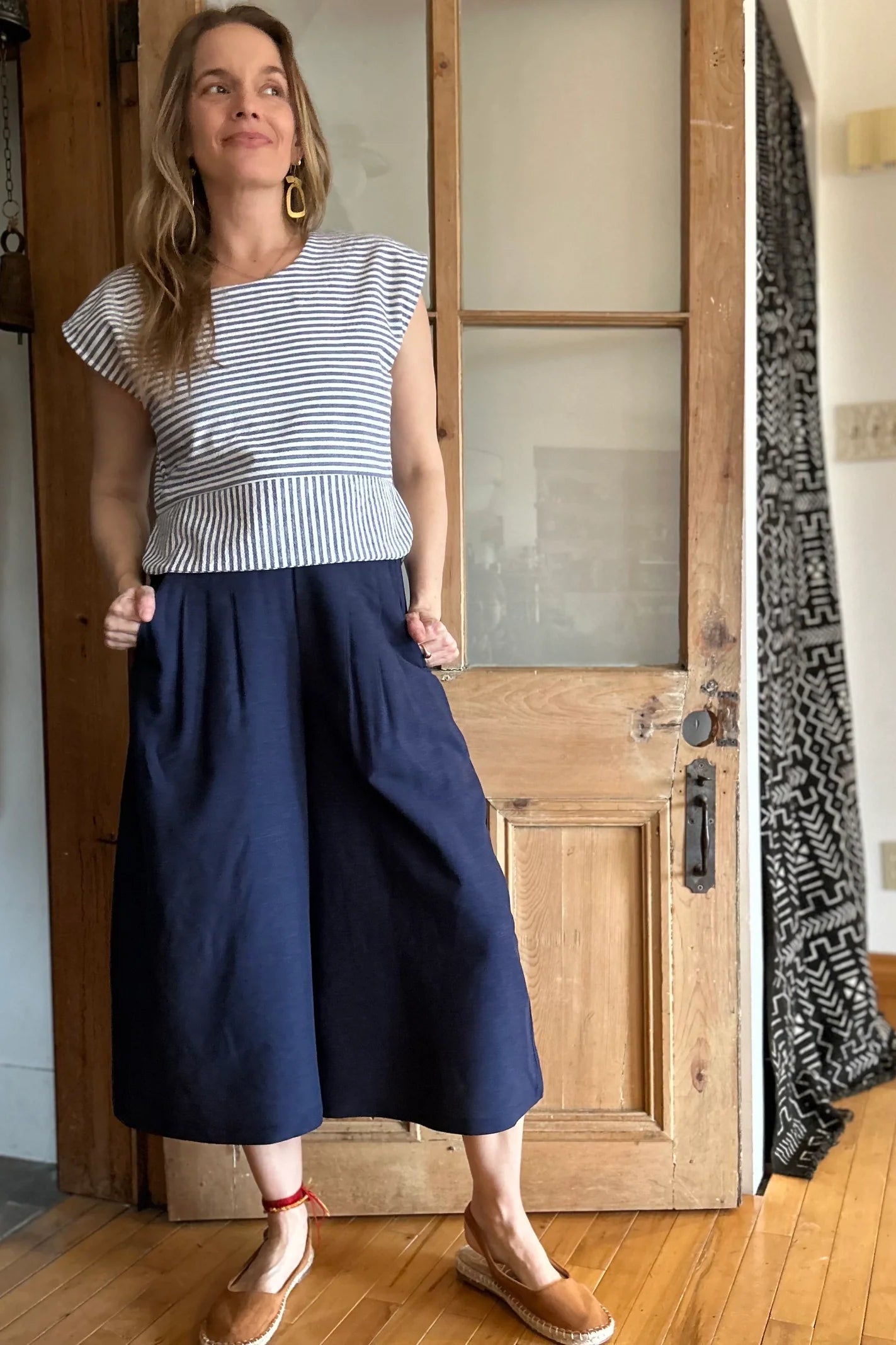 A woman wearing the Petunia Pants by Kazak in Indigo, wide legged cropped pants with a waistband that's smooth in front but elastic at the back, front pleats, and pockets. She is wearing them with the Tulipe top and standing in front of a wooden door. 