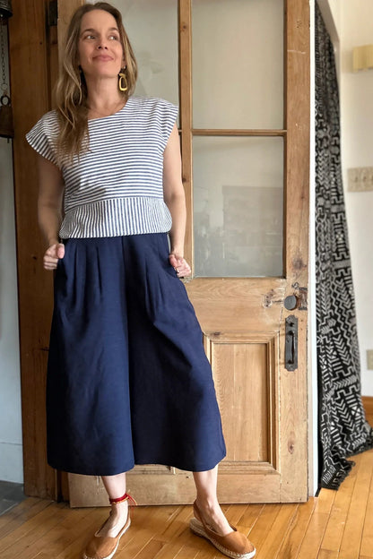 A woman wearing the Petunia Pants by Kazak in Indigo, wide legged cropped pants with a waistband that's smooth in front but elastic at the back, front pleats, and pockets. She is wearing them with the Tulipe top and standing in front of a wooden door. 