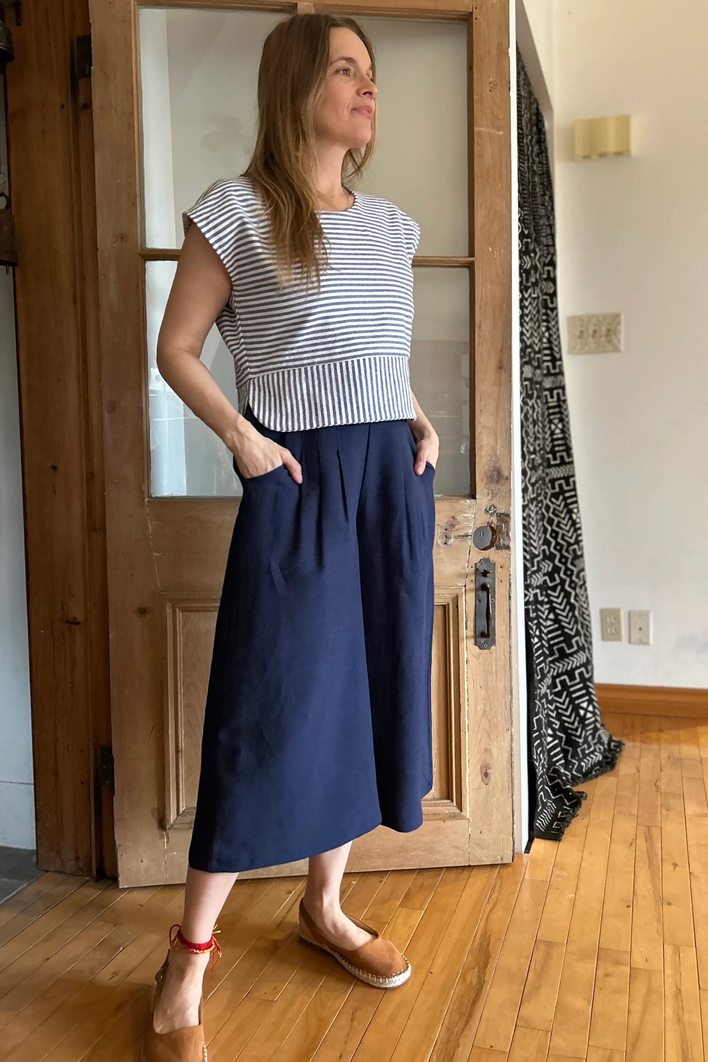 A woman wearing the Petunia Pants by Kazak in Indigo, wide legged cropped pants with a waistband that's smooth in front but elastic at the back, front pleats, and pockets. She is wearing them with the Tulipe top and standing in front of a wooden door. 