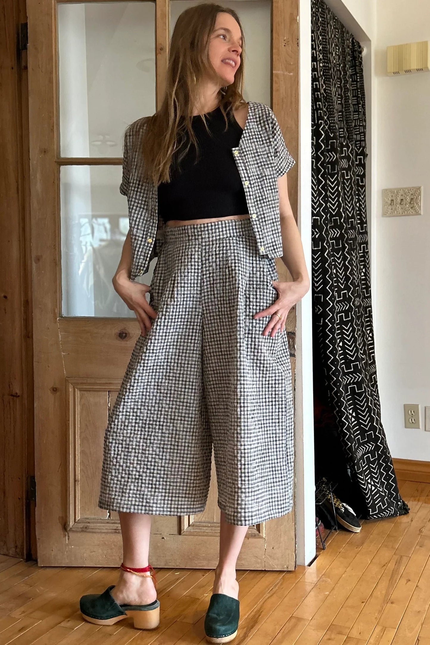 A woman wearing the Petunia Pants by Kazak in Black and Cream Vichy/Gingham, wide legged cropped pants with a waistband that's smooth in front but elastic at the back, front pleats, and pockets. She is wearing them with a black tank and matching top and standing in front of a wooden door. 