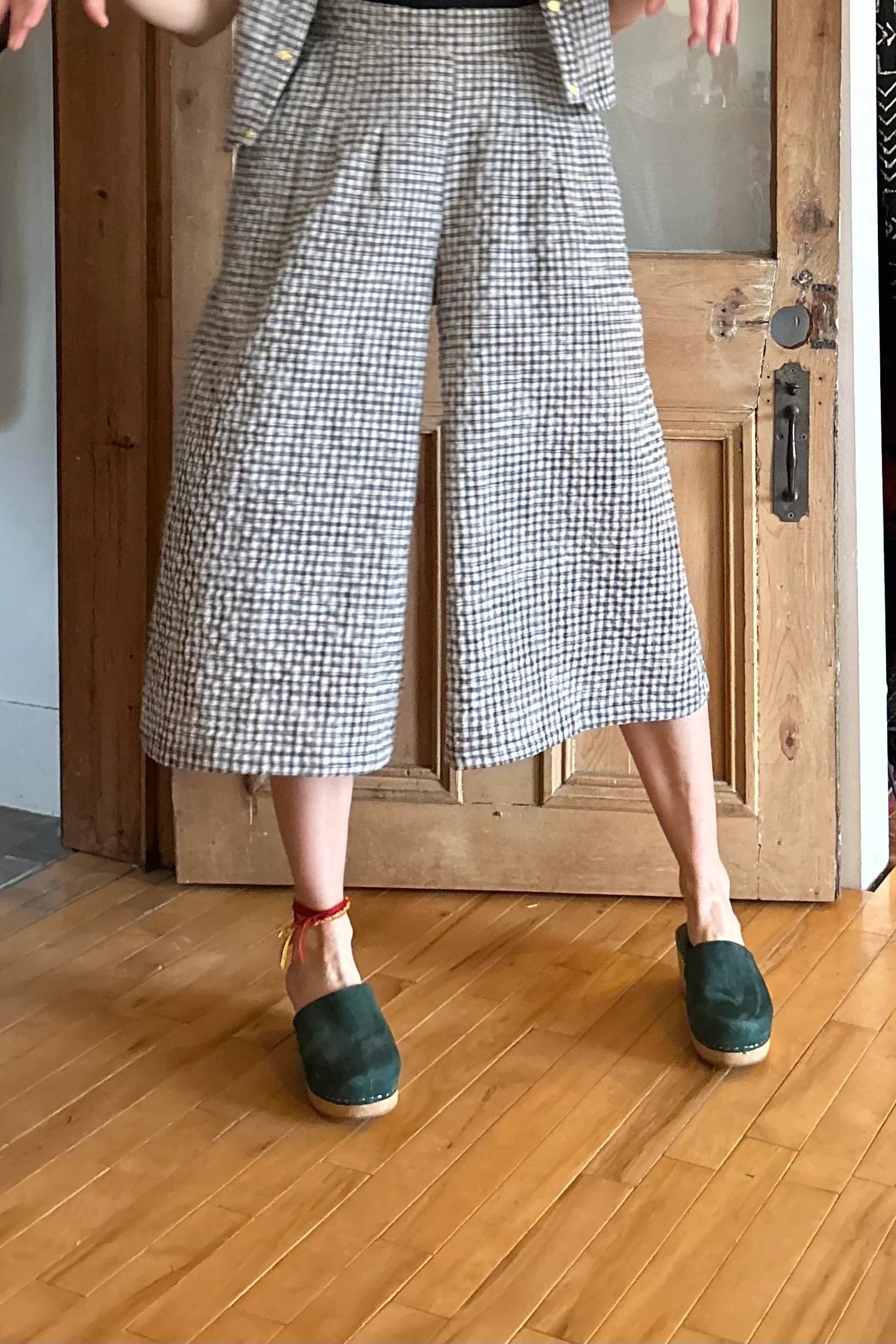 Waist down view of a woman wearing the Petunia Pants by Kazak in Black and Cream Vichy/Gingham, wide legged cropped pants with a waistband that's smooth in front but elastic at the back, front pleats, and pockets. She is wearing them with a black tank and matching top and standing in front of a wooden door. 