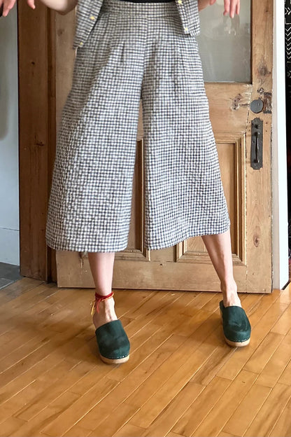 Waist down view of a woman wearing the Petunia Pants by Kazak in Black and Cream Vichy/Gingham, wide legged cropped pants with a waistband that's smooth in front but elastic at the back, front pleats, and pockets. She is wearing them with a black tank and matching top and standing in front of a wooden door. 