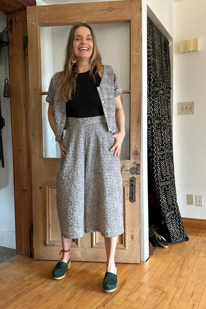 A woman wearing the Petunia Pants by Kazak in Black and Cream Vichy/Gingham, wide legged cropped pants with a waistband that's smooth in front but elastic at the back, front pleats, and pockets. She is wearing them with a black tank and matching top and standing in front of a wooden door. 