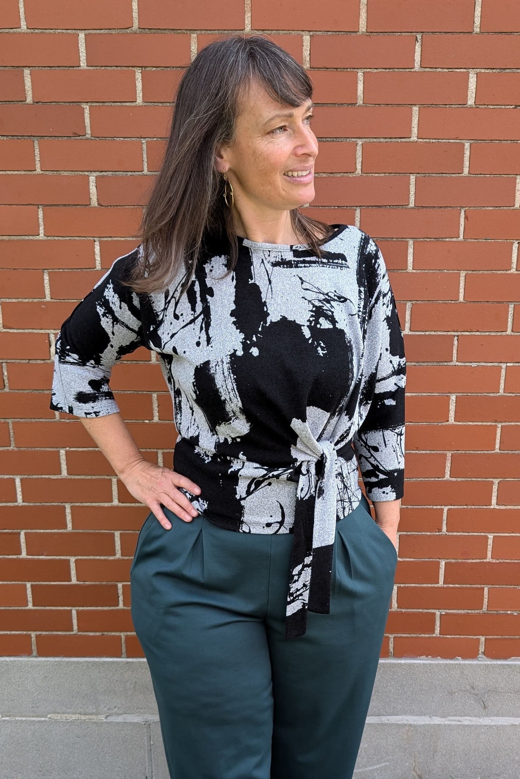 A woman wearing the NIna Top by Compli K, a knit top in a grey and black abstract pattern with 3/4 sleeves and a tie detail at the front, with the Wilfred Pants by Melow in Forest, standing in front of a brick wall. 