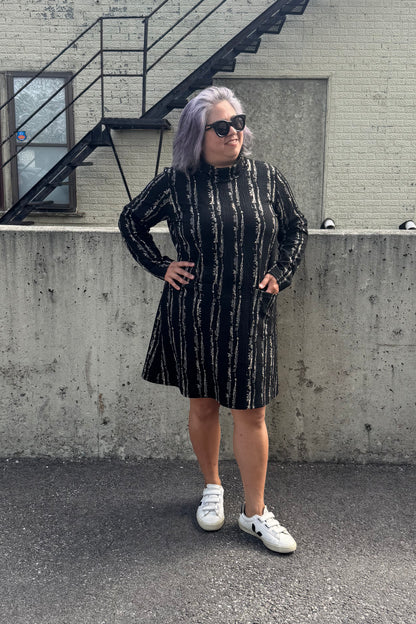 A woman wearing the Floella Dress by Pure Essence in Black/Tan, featuring a turtleneck, long sleeves, a slight a-line shape that hits the knee, and front patch pockets. She is standing in front of a grey wall. 