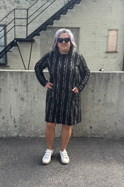 A woman wearing the Floella Dress by Pure Essence in Black/Tan, featuring a turtleneck, long sleeves, a slight a-line shape that hits the knee, and front patch pockets. She is standing in front of a grey wall. 