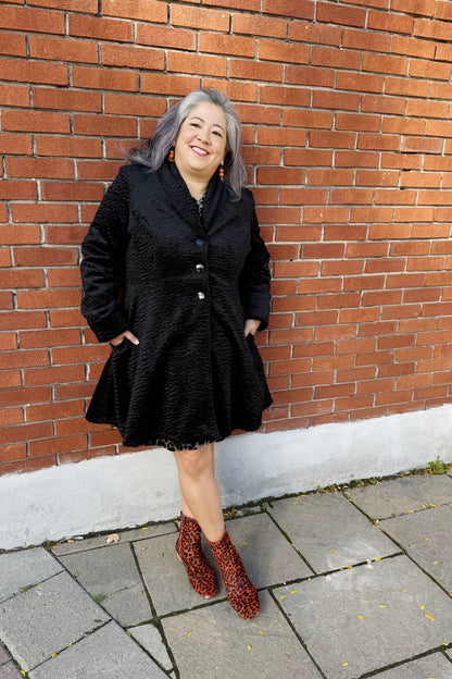 A woman wearing the Spies Jacket by Luc Fontaine,  
a fit and flare above the knee coat in boucle fabric, standing in front of a brick wall 
