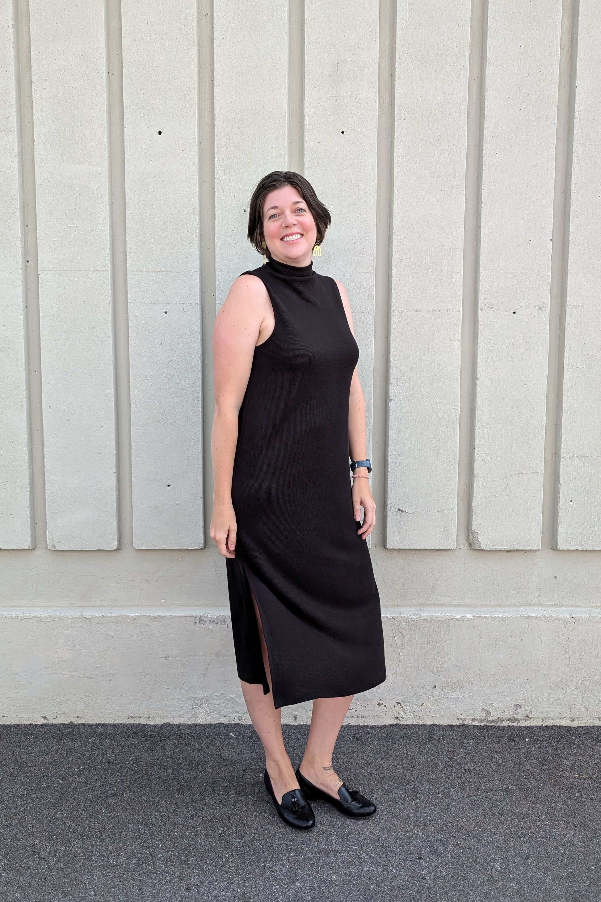 A woman wearing the Natasha Dress by Compli K in Black, a sleeveless midi-length dress with a mock turtleneck and side slits, standing outside in front of a white wall 