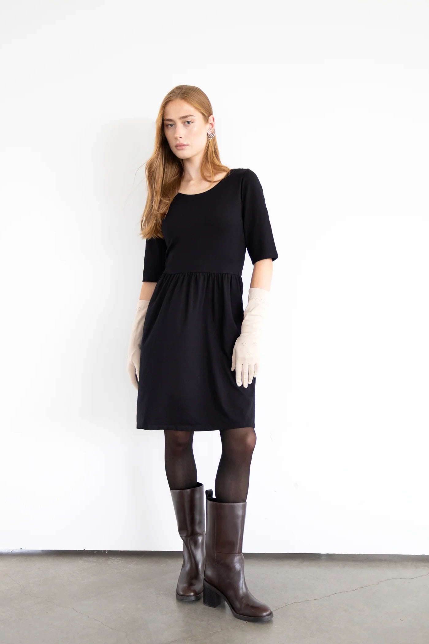 A woman wearing the Smith Dress by Eve Lavoie in Sparkly Black, a reversible dress with a round neck one one side, v-neck on the other, elbow length sleeves, and knee-length A-line skirt. She is standing in front of a white background. 