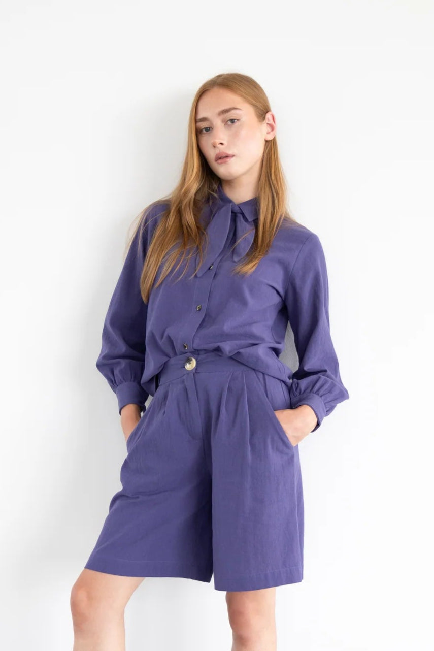  A woman wearing the Blair Shirt by Eve Lavoie in Violet, a button front shirt with a removable neck tie, rounded hem, and puffed sleeves. She is wearing it with matching shorts and standing in front of a white background. 