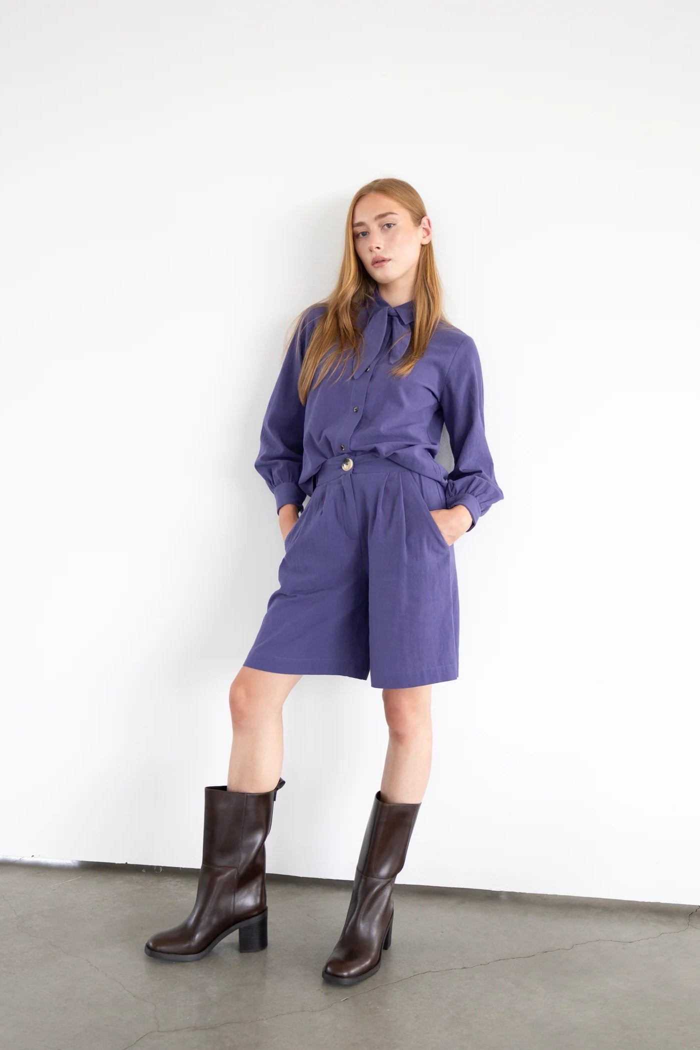  A woman wearing the Blair Shirt by Eve Lavoie in Violet, a button front shirt with a removable neck tie, rounded hem, and puffed sleeves. She is wearing it with matching shorts and standing in front of a white background. 